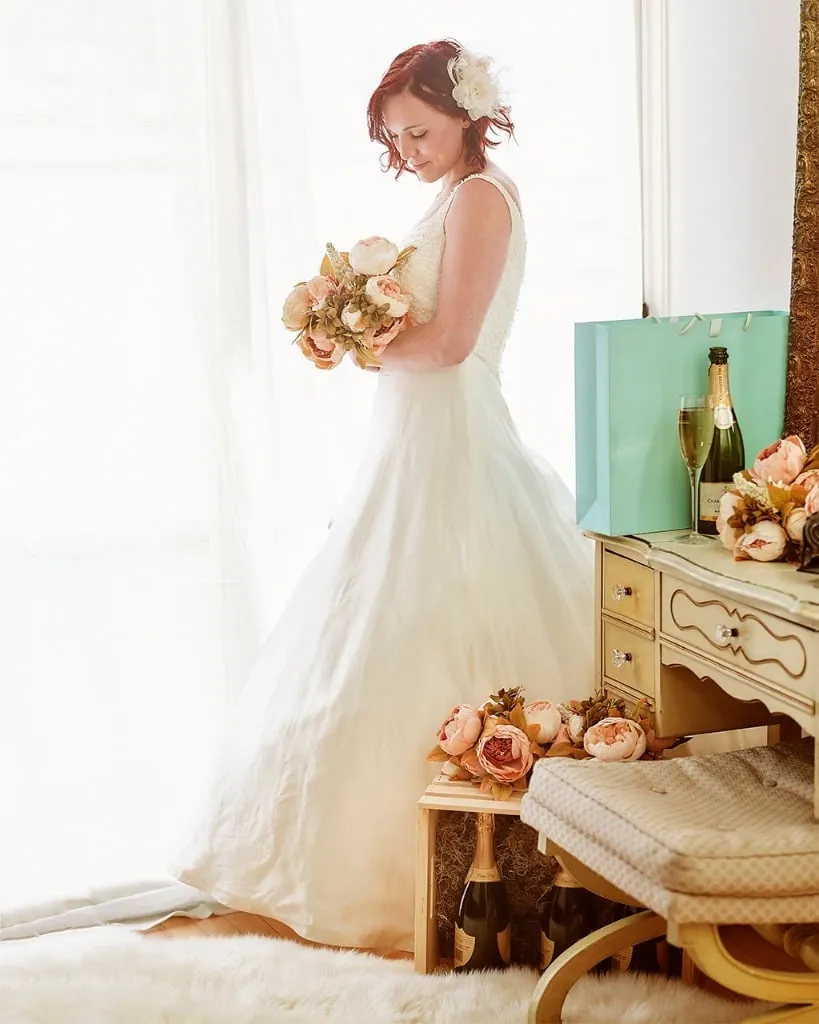 Brightly lit bridal portrait of red haired woman holding a bouquet facing towards a curtained window