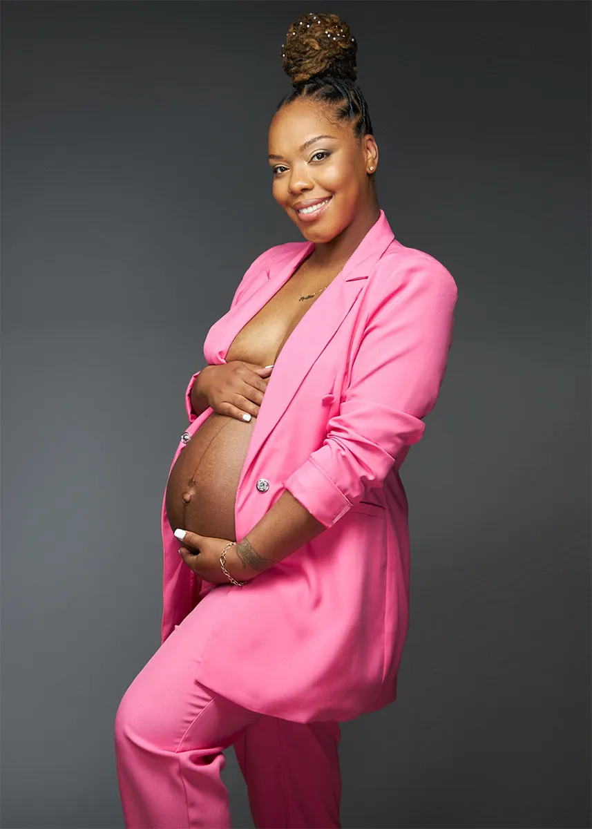 Maternity portrait of woman in a pink pantsuit posed in front of a grey backdrop