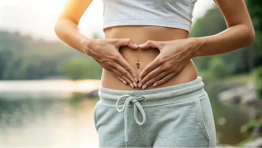 a woman's midsection, with her hands forming a heart shape over their stomach. The person is wearing light gray, casual sweatpants with a drawstring. The background is blurred and natural, suggesting an outdoor or serene environment. The image symbolizes gut health, self-care, and a focus on well-being.