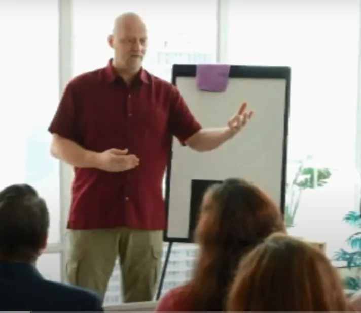 Chad Nedland speaking in front of a classroom durin a mastermind wearing a maroon dressshirt and tan kahki pants