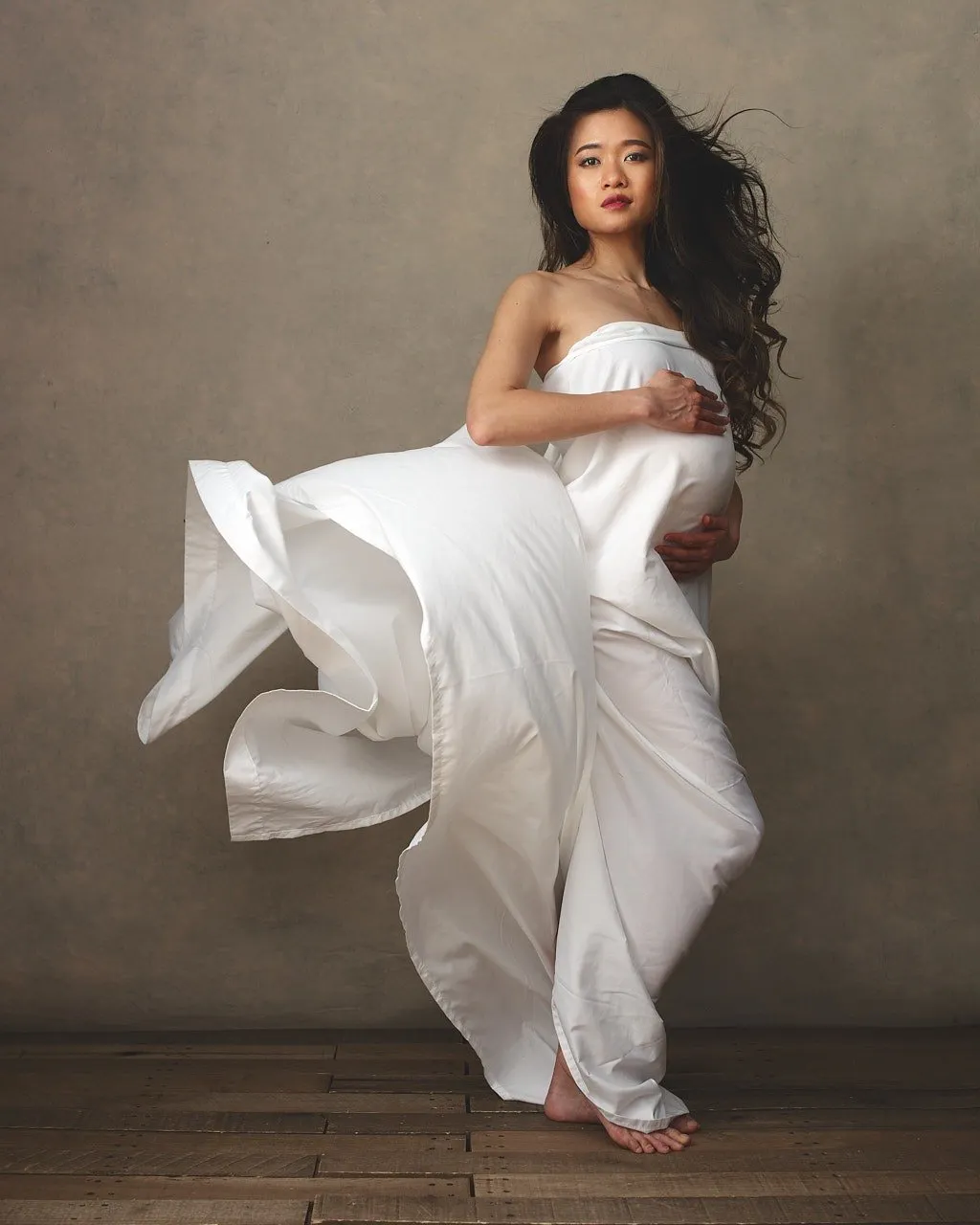 Neutral toned maternity portrait of Asian woman covered by a white sheet posed in front of a taupe background