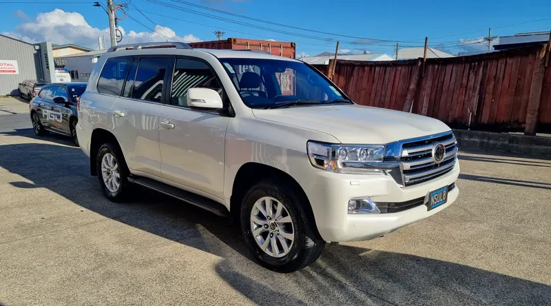 A white Toyota Land Cruiser SUV is parked on a sunlit concrete surface in an industrial area. The vehicle features chrome accents, silver alloy wheels, and tinted windows. In the background, there are other vehicles, including a black car with 