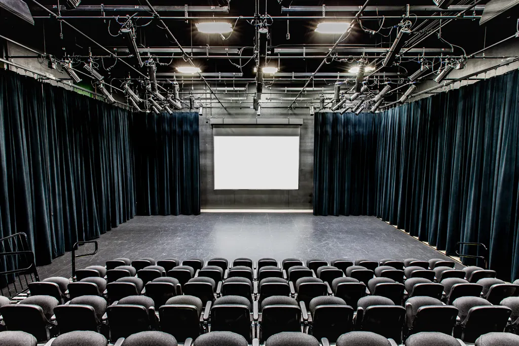 Interior view of a black box theatre featuring a minimalist stage, black curtains on all sides, ceiling-mounted lighting equipment, and rows of black chairs facing the stage. A projection screen is centered at the back of the stage.