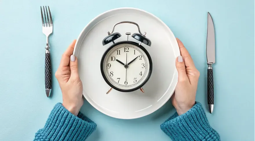 a white plate with a classic black alarm clock placed in the center, symbolizing meal timing. The clock's hands indicate a specific time, reinforcing the concept of structured eating.