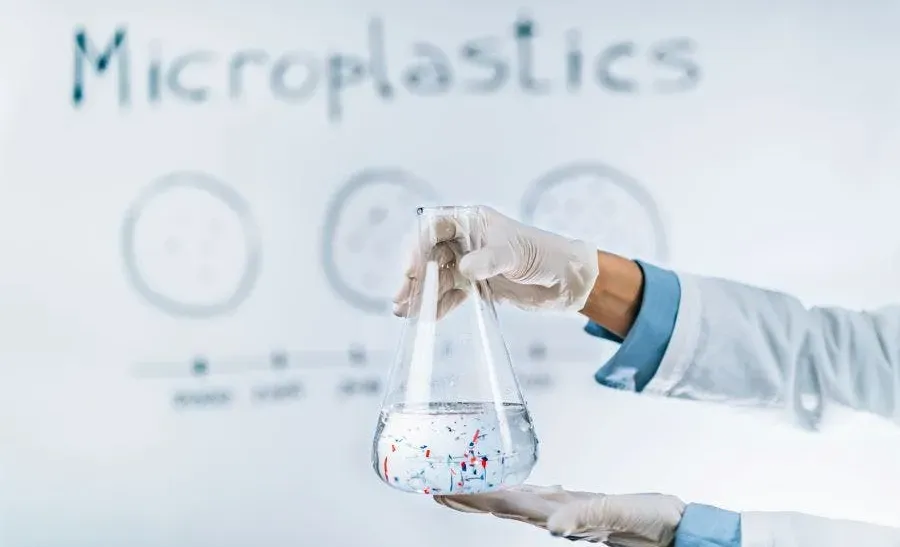 a scientist holding a large flask filled with water containing visible microplastic particles. The scientist is wearing white gloves, and in the background, the word "Microplastics" is visible on a whiteboard