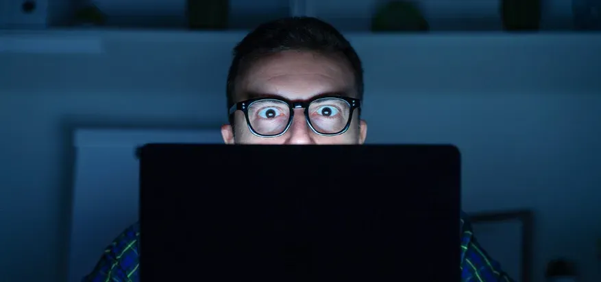 Man looking at his computer screen in the dark with blue light illuminating the room.