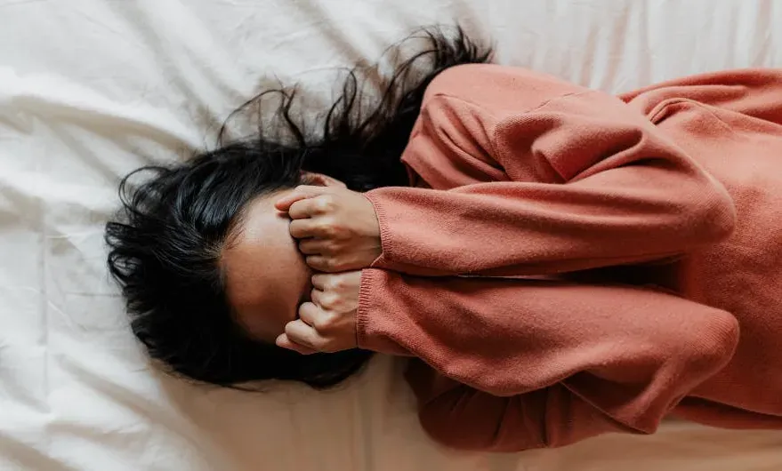 A woman lying on her bed with her hands covering her face appearing to be distressed. 