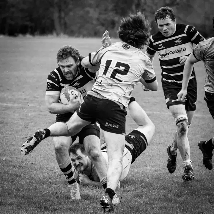 Black and white rugby photograph showing a ball carried straight arming a defender while another defender who missed his tackle is in the background