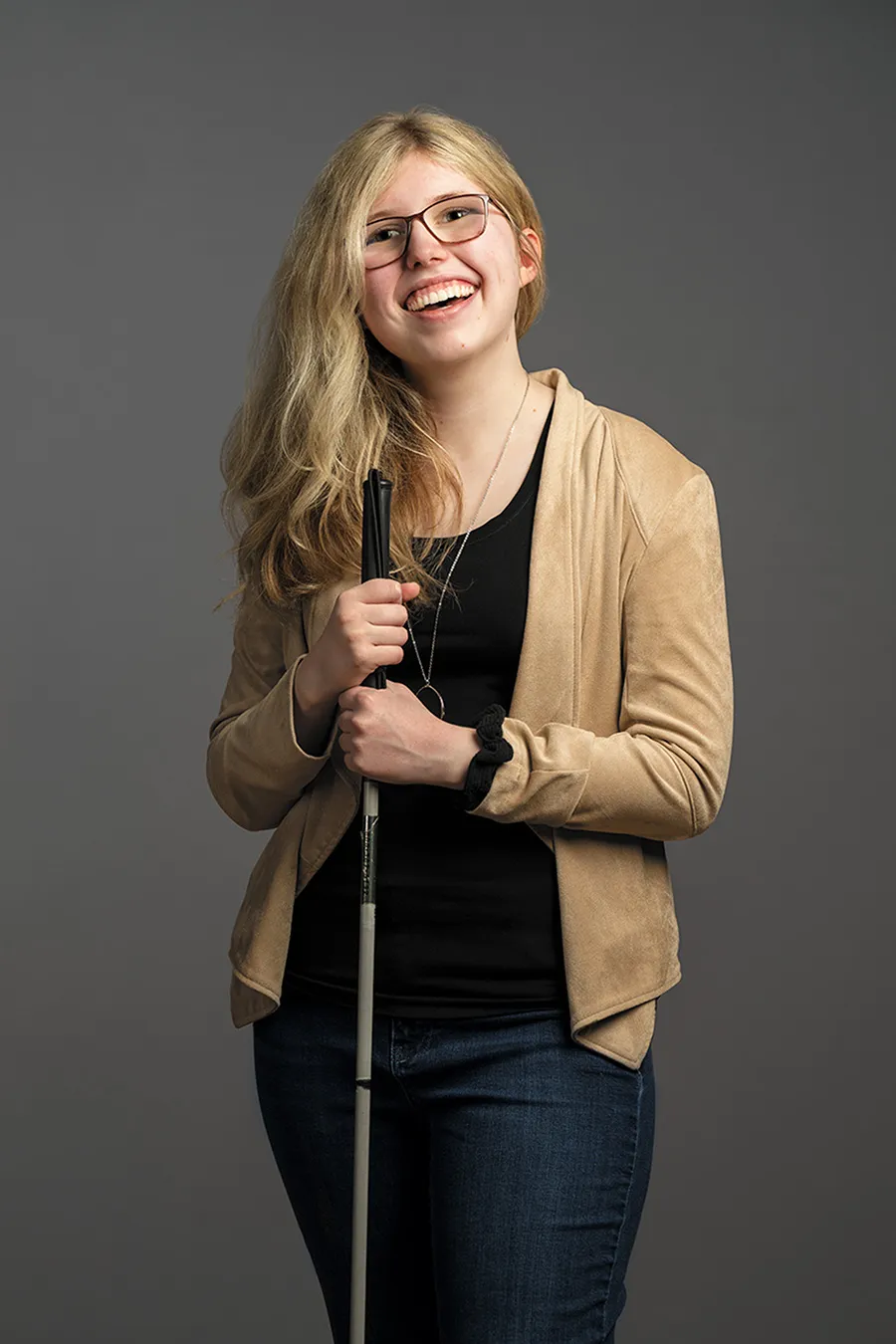 kaleigh with long blonde hair smiling wearing a tan blazer and dark pants holding her white cane