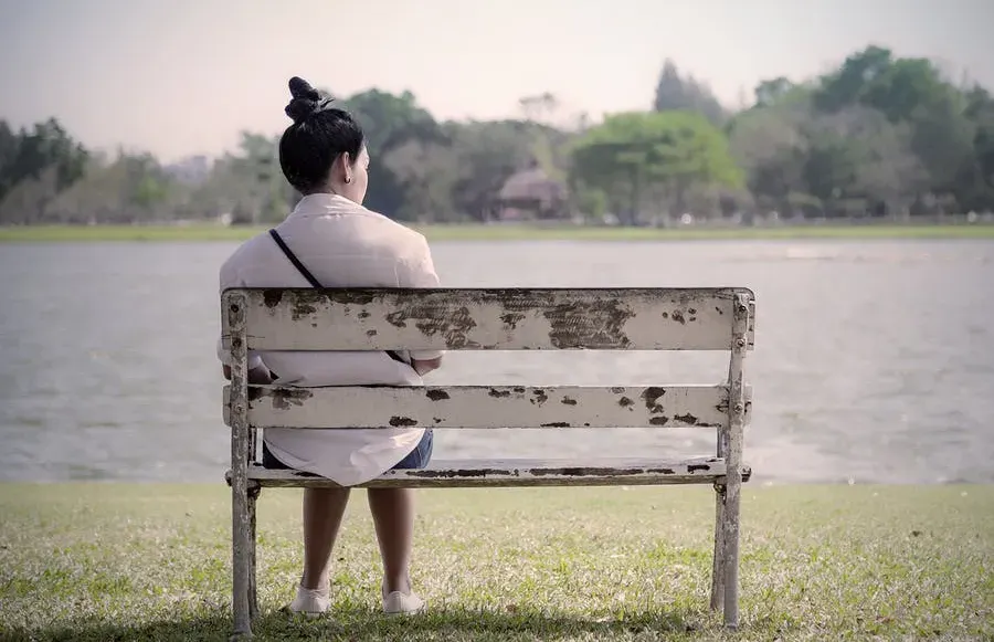 A woman sitting alone by herself on an old, worn-out park bench by a lake.