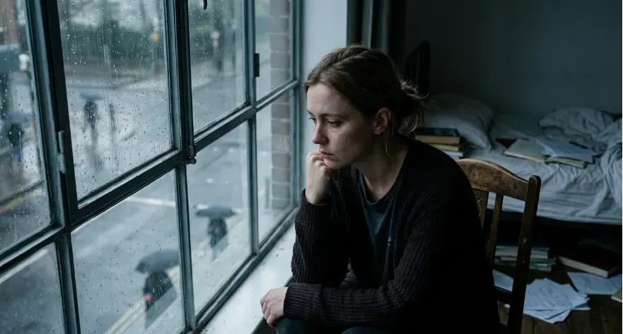 Person sitting alone in a dim room staring out the window on a rainy, gloomy day looking sad and thoughtful