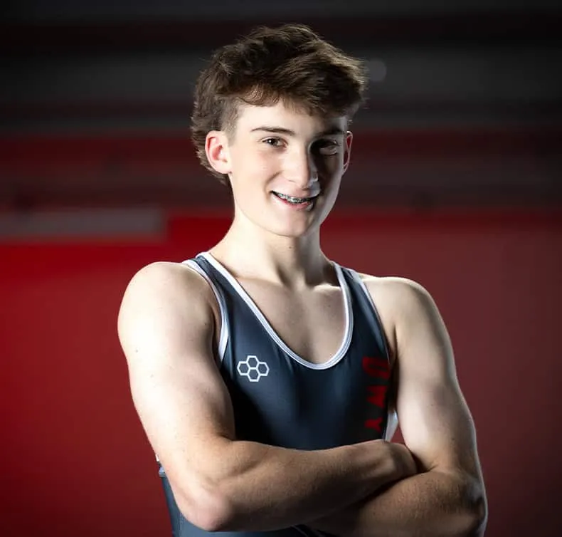 Young Owyhee Storm high school wrestler smiling confidently with braces, wearing navy blue and white wrestling singlet, arms crossed in professional sports portrait against dramatic red background