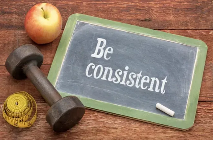 A small chalkboard with the words "Be Consistent". It's surrounded by a dumbbell, measuring tape, and an apple