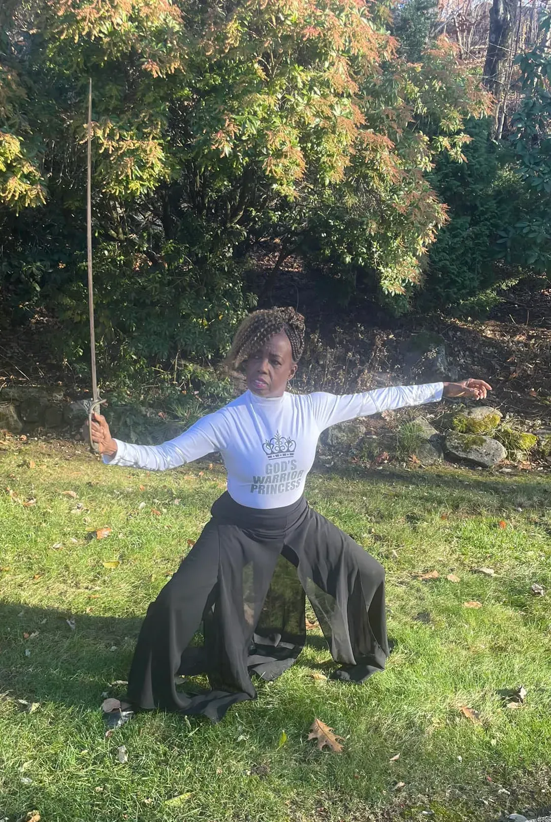 Coach Judy Handwerker performs a graceful worship-inspired movement outdoors while holding a sword in a low lunge position. Dressed in a white 