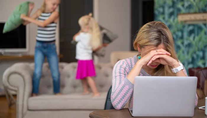 Mom stressed at laptop with kids playing in background.