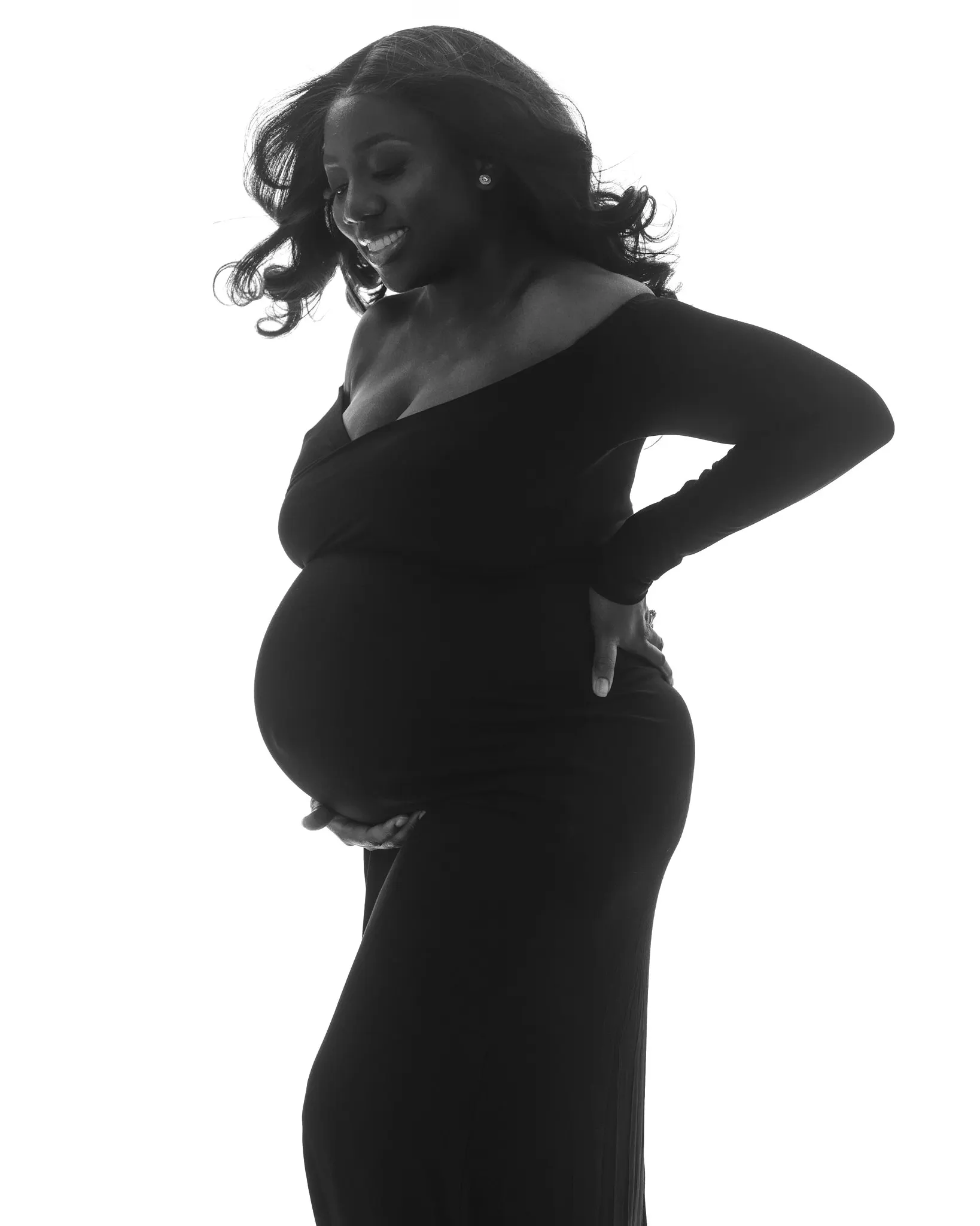 Darkly lit maternity portrait of woman in a black turtleneck and jeans posed in front of a dark grey backdrop